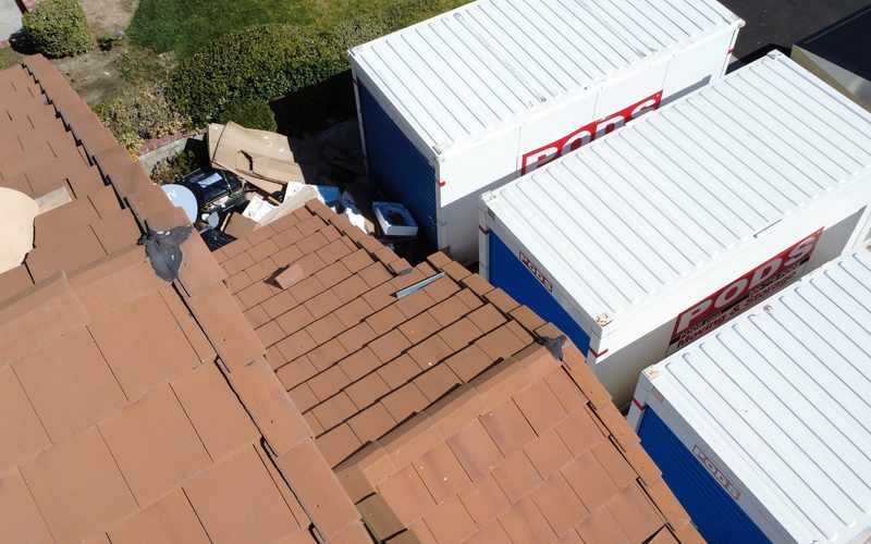 Aerial view of terracotta tile roof planes, ridges, and roof penetrations, with portable storage containers standing in the driveway beyond.