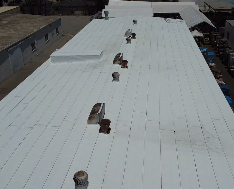 Aerial view of a long white coated low-slope roof with evenly spaced parallel seams and a row of round metal vents down the center.