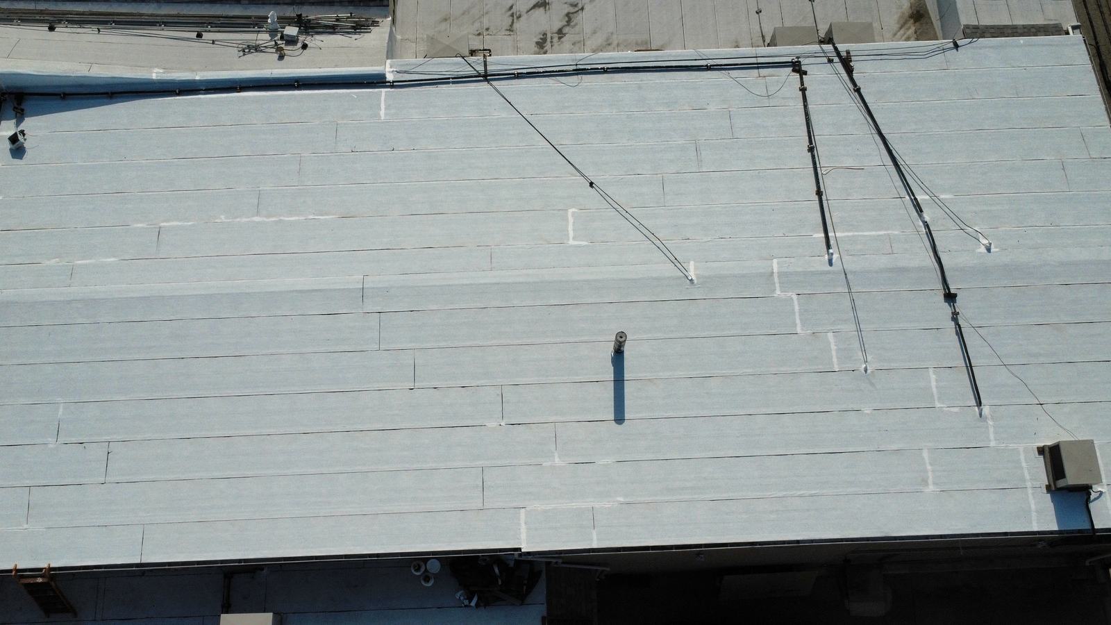 Straight-down aerial of a complete commercial low-slope roof: a broad white-coated membrane field with visible seam lines, parapet and drip edge on three sides, a vent stack, a rooftop mechanical unit, conduit runs and a neighboring roof deck.