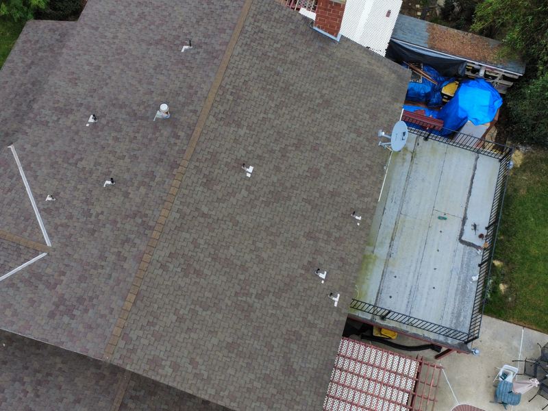 High-angle aerial of a weathered gray-brown asphalt shingle roof plane with a hip at left, small white roof covers, an adjacent corrugated metal patio roof, a blue drum and a red tile paved area below.