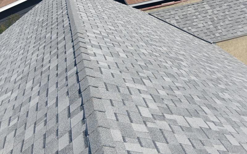 Elevated view over a finished gray architectural-shingle roof: a large hip plane with ridge and hip caps in the foreground, a second shingle roof section and a stucco wall behind, plus nearby residential roof context.