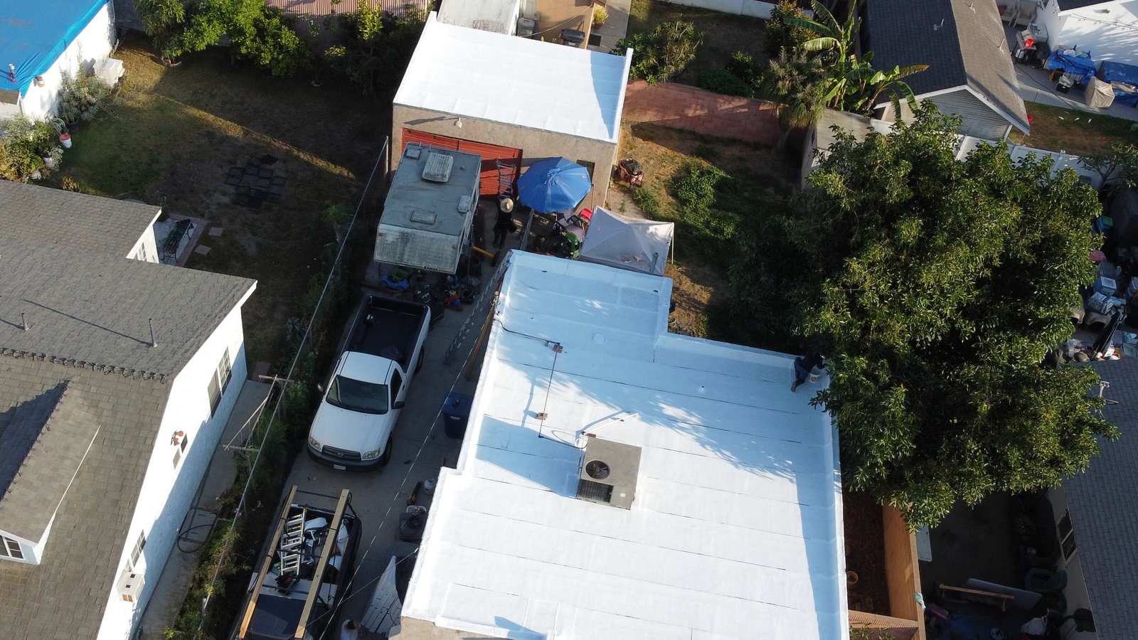 Aerial of an active residential low-slope job: a white-coated membrane roof filling the center of the frame with a rooftop penetration and curb, a person at the roof edge, another person at ground level, a ladder, staged materials and work trucks in the driveway.