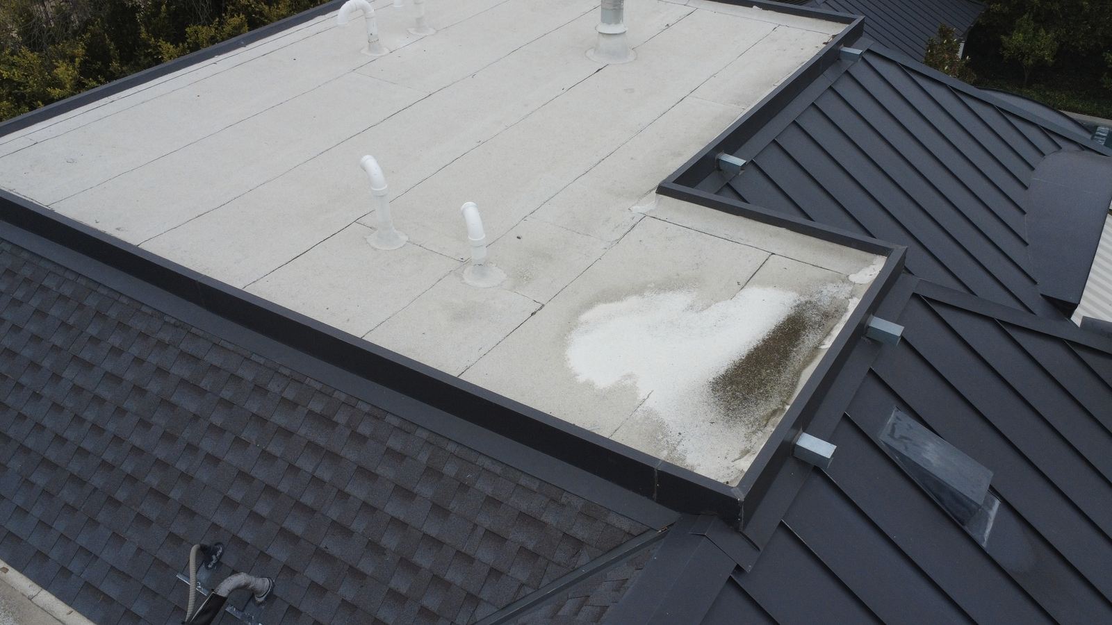 High aerial of a complete multi-system residential roof: a parapet-bounded white low-slope field with five plumbing vents and a flue, adjacent finished dark standing-seam metal planes with hips, ridge caps and a curb-mounted skylight, and an architectural shingle plane at lower left.