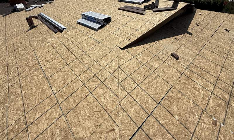 A large bare OSB roof deck spanning several planes with visible panel seams and fastener lines, two framed skylight curbs and a hip meeting the ridge, with staged material on the deck under a clear blue sky.