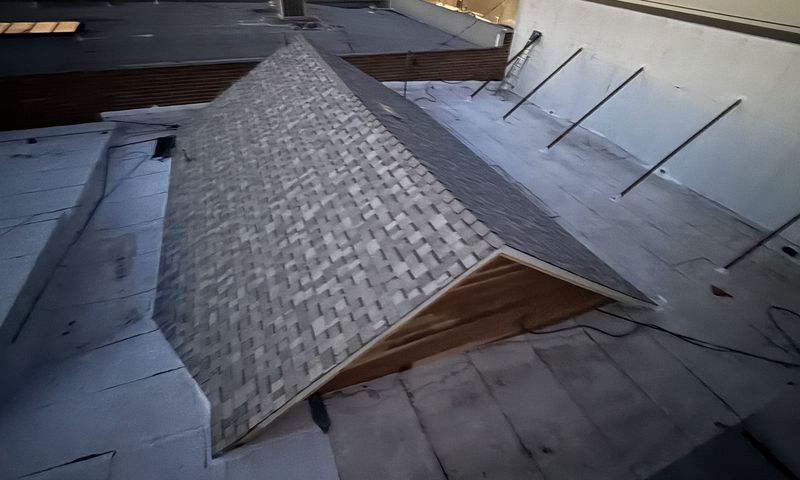 A dark gray asphalt shingle hip roof section rising out of a surrounding flat low-slope roof area bounded by light stucco parapet walls, with a metal railing along the upper edge and an adjacent building wall closing the background, under overcast daylight.