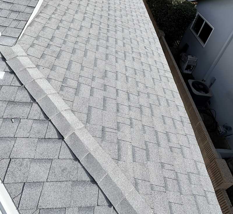 A light-gray asphalt shingle hip roof photographed from roof level, with a hip line running down the center of the frame, ridge caps along the top edge, and a light stucco house wall and garden foliage at the right.