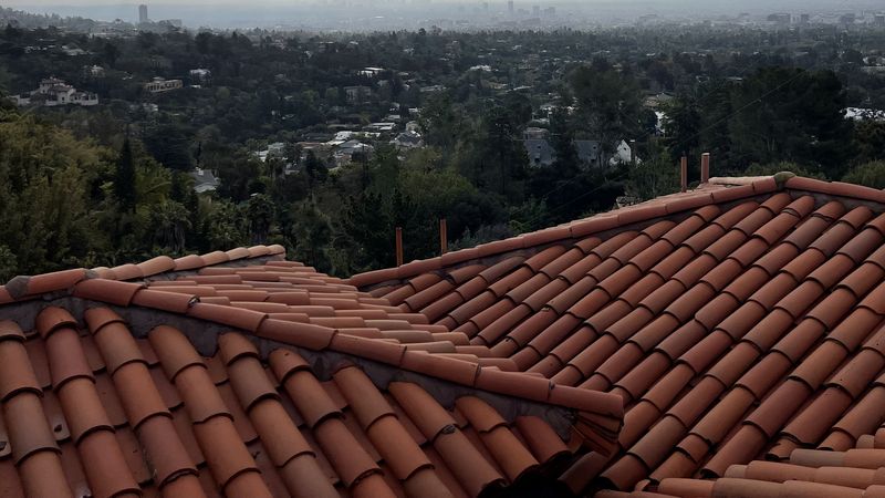Red barrel-tile roof planes and a ridge running across the foreground with a thin strip of hazy valley horizon beyond, under a heavily clouded sky.