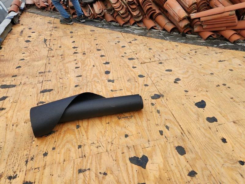 Complete tile re-roof job stage: a full stripped plywood deck plane with old nail and bitumen residue, salvaged clay barrel tiles stacked in long rows along the upper edge, a partially unrolled underlayment roll on the deck, and a worker's lower legs and boots at the top edge.